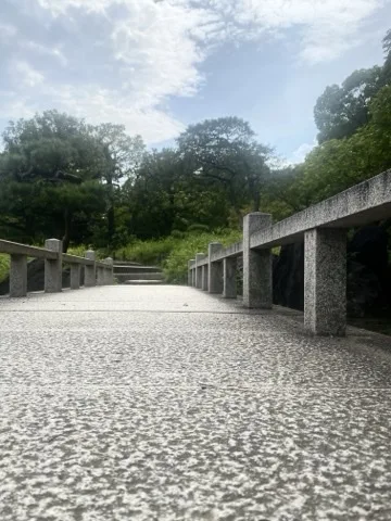 石橋の路面すれすれから見た景色。左右に低い石の欄干が続き、その先に石段と木々、空。
