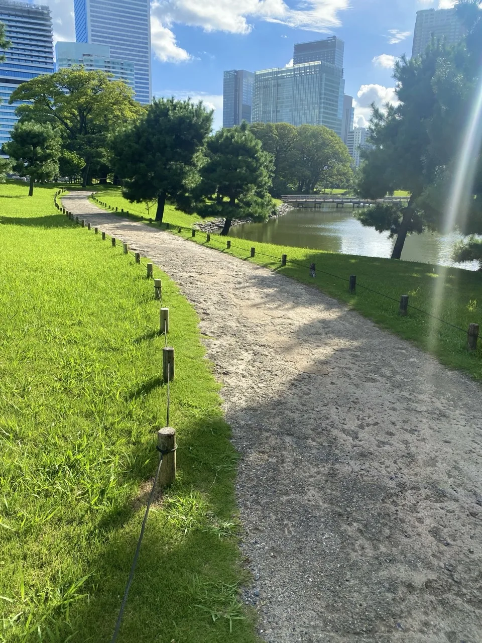 白い砂利道が芝の間を緩く曲がって続く。右手に堀の水面、奥に木立と高層ビル。