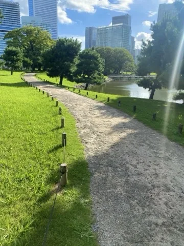 白い砂利道が芝の間を緩く曲がって続く。右手に堀の水面、奥に木立と高層ビル。
