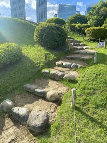 芝の斜面を上る土の段。各段の縁に丸い石が並び、両側に刈り込みの低木と縄の柵。奥に高層ビル。
