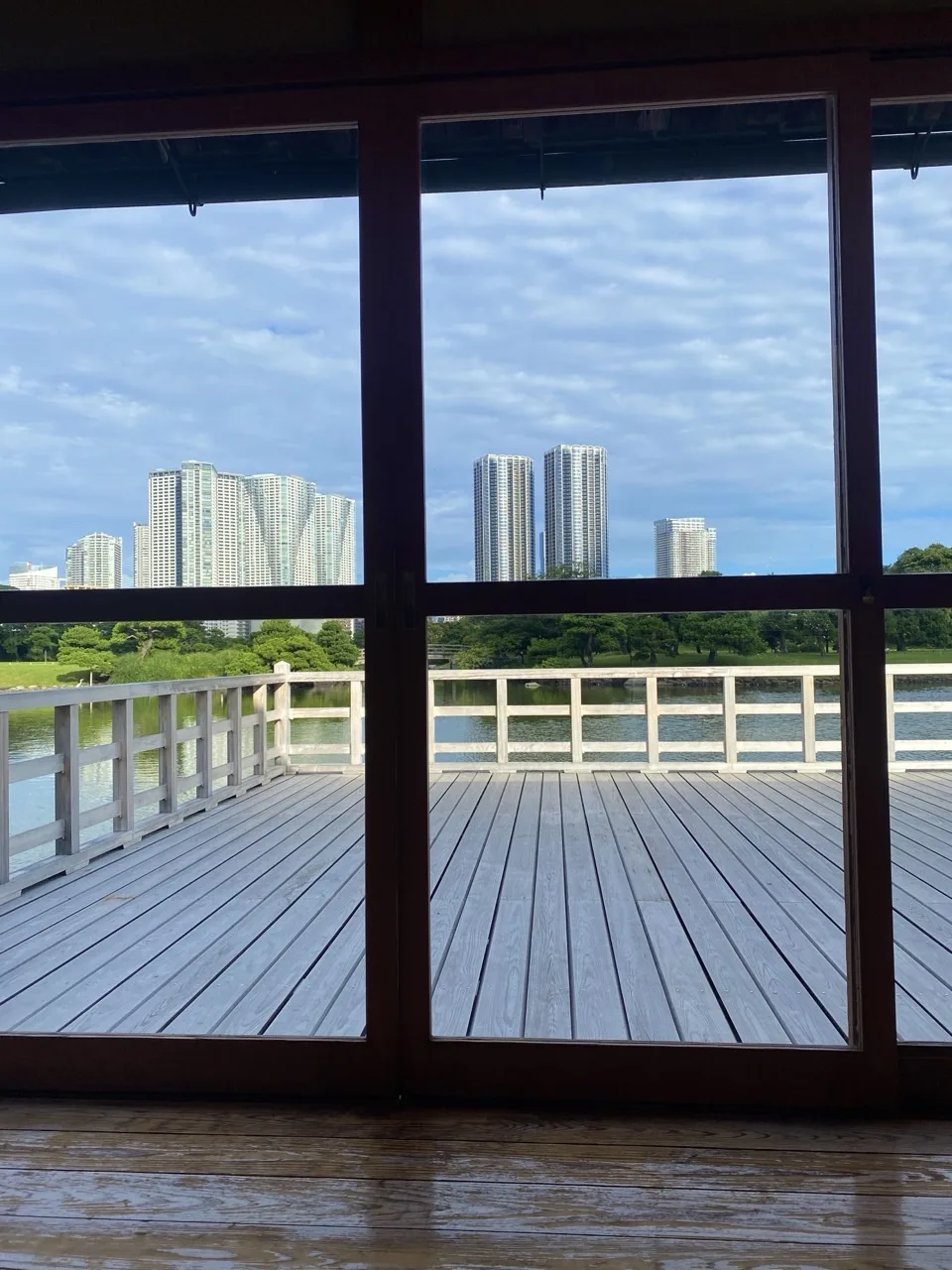 建物の内側から見た開口。太い柱と桟が眺めを分け、その向こうに白い欄干の縁側、池、対岸の緑と高層ビル。