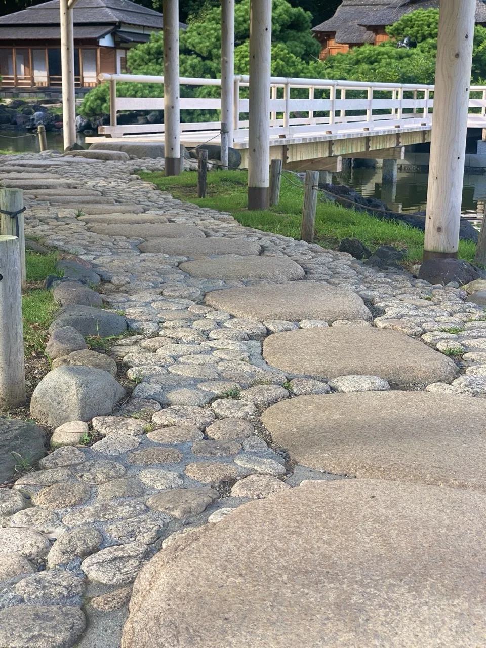 大きな平らな石と丸い小石を組み合わせた道が奥へ続き、その先に白い欄干の橋と茅葺きの建物がある。