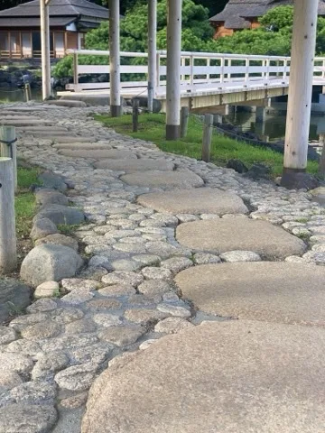 大きな平らな石と丸い小石を組み合わせた道が奥へ続き、その先に白い欄干の橋と茅葺きの建物がある。