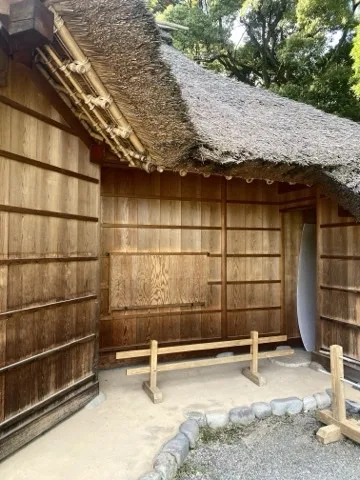 板壁と茅葺き屋根の建物の軒下に、細い木の腰掛けが一つ置かれている。足元は砂利で、右手に丸い石が並ぶ。