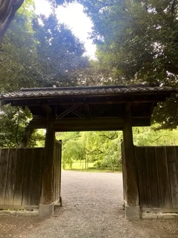 茅葺屋根の木の門を正面から見た写真。奥に砂利の園路と木立が続く。