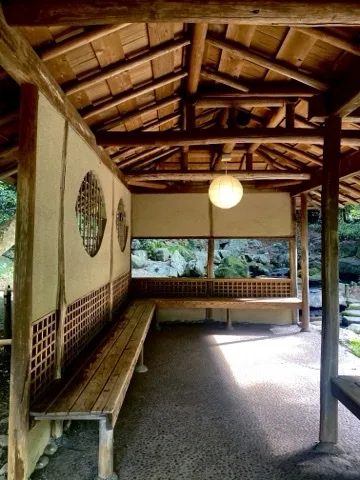 茅葺屋根の東屋の内部から、柱の間に庭園の緑を見た写真。行灯が灯る。