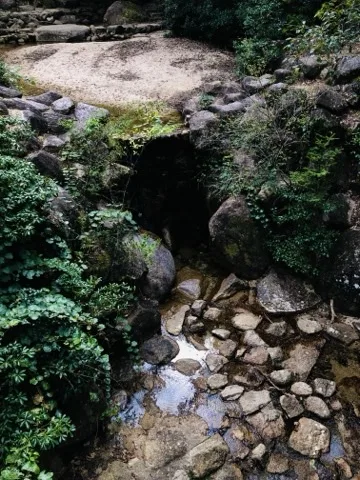 石を組んだ谷筋の下に暗い開口があり、そこから浅い流れが手前へ続く。周りを苔と低木が覆う。