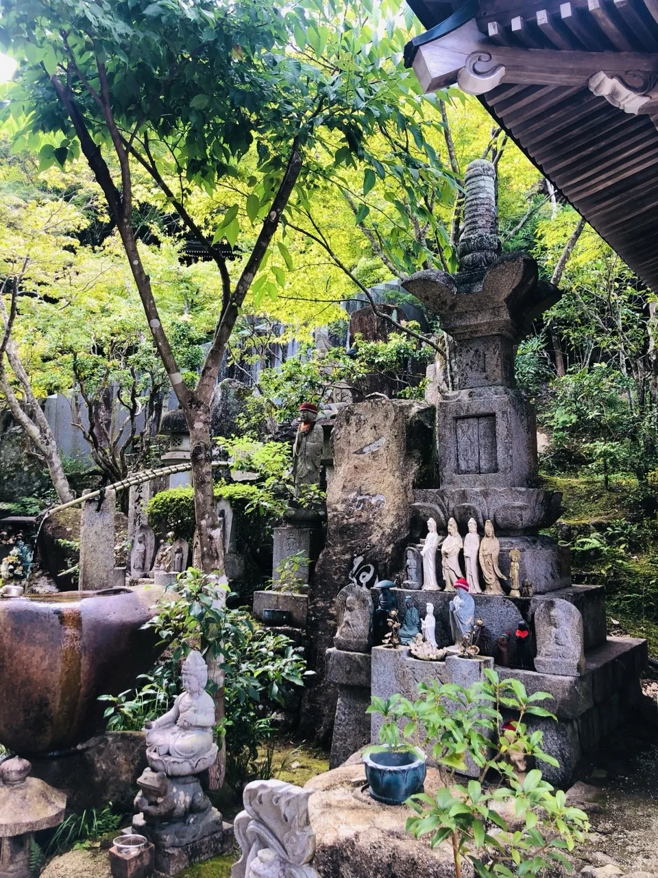 木立の下の斜面に石塔と小さな石像が幾つも並び、その手前に苔むした据え石と鉢がある。右上に建物の軒。