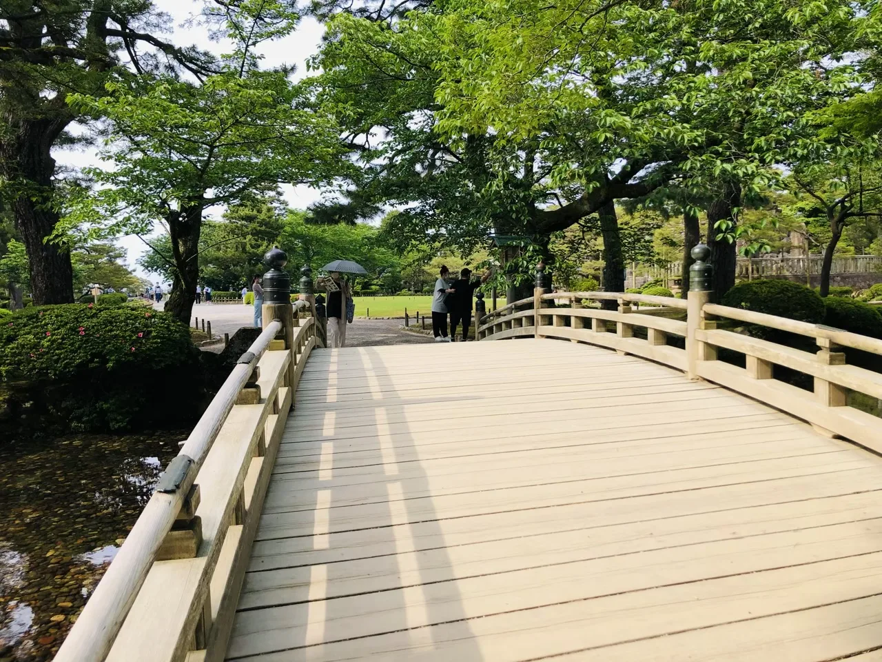 白木の反り橋の上から見た画面。欄干が左右にあり、渡り終えた先に木立と芝地、数人の人影がある。