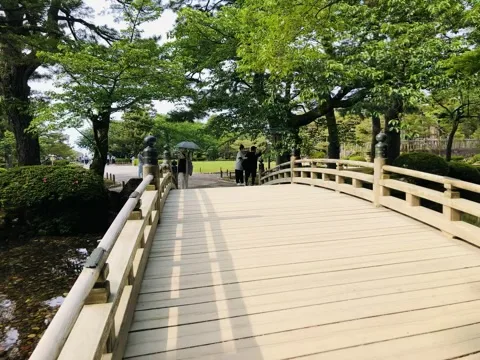 白木の反り橋の上から見た画面。欄干が左右にあり、渡り終えた先に木立と芝地、数人の人影がある。
