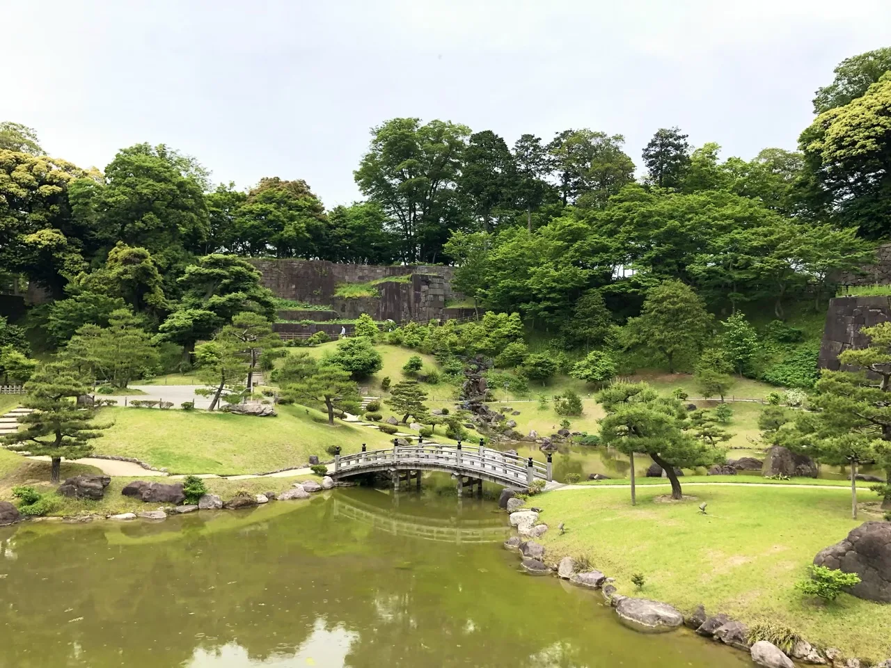 手前に緑色の水をたたえた池、中央に白い反り橋が架かる。背後の斜面は芝と刈込みの松、その上に城の石垣が段を成して続く。