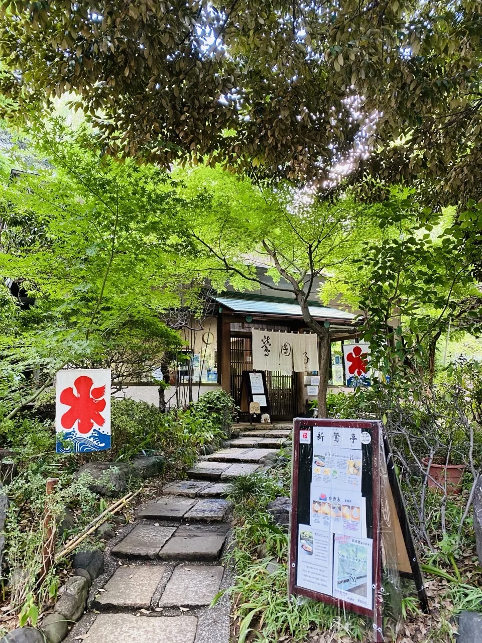 石を敷いた段が奥の小さな建物へ上っていく。左右に赤い「氷」の旗と品書きの立て看板があり、周囲は明るい緑の枝葉に囲まれている。