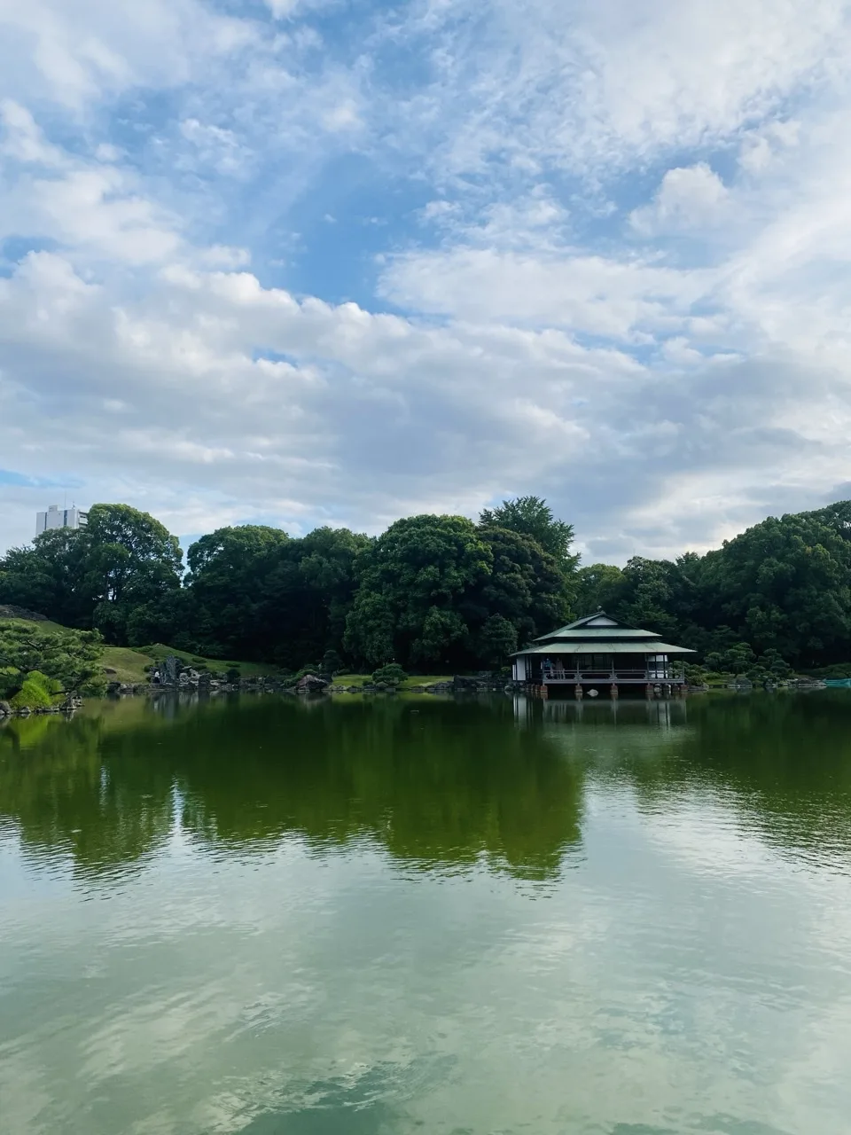 静かな池の向こう岸に、銅葺き屋根の水辺の建物が張り出して建っている。周囲は濃い緑の常緑樹に囲まれ、空には白い雲が広がり、その姿が緑色の水面に映っている。左奥にビルの上部がわずかに見える