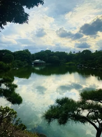 画面の上部と下部を木の枝と松の葉が縁取り、その間から広い池が見える。対岸中央に瓦屋根の水辺の建物が建ち、静かな水面には夕方の白い雲がくっきりと映り込んでいる