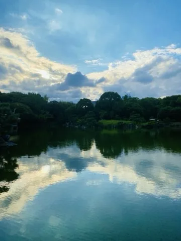 夕方の池。空には日の光を含んだ雲が広がり、その明るい姿が静かな水面いっぱいに映り込んでいる。対岸の木立は逆光で暗いシルエットになり、右手に草地の斜面が見える。左端に白い鳥の姿がある