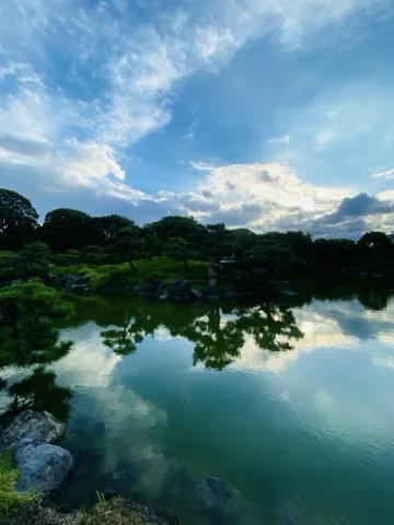 夕暮れ近い池の風景。対岸の岩で護岸された岸に背の高い石灯籠が立ち、その周りに丸く刈り込まれた松や草地が広がる。手前左には松の枝と大きな岩があり、水面は青い空と雲を映して穏やかに光っている