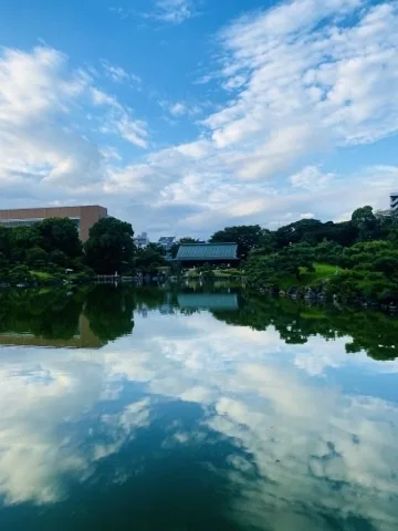 波のない池の水面いっぱいに青空と白い雲が映っている。対岸中央には緑青色の大屋根をもつ和風の建物があり、その左右に木立と刈り込まれた植栽、右手には岩組みの護岸が続く。左奥には大きなビルが見える