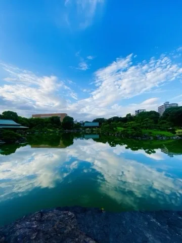 広い池の水面に青空と白い雲がそのまま映る。両岸に木立と建物、手前に黒い岩。