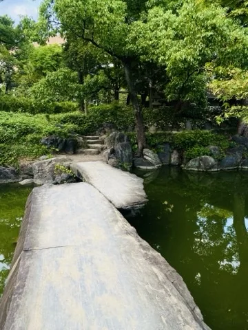池に架かる細長い一枚石の橋。渡った先に石段と木立、右手に緑色の水面。