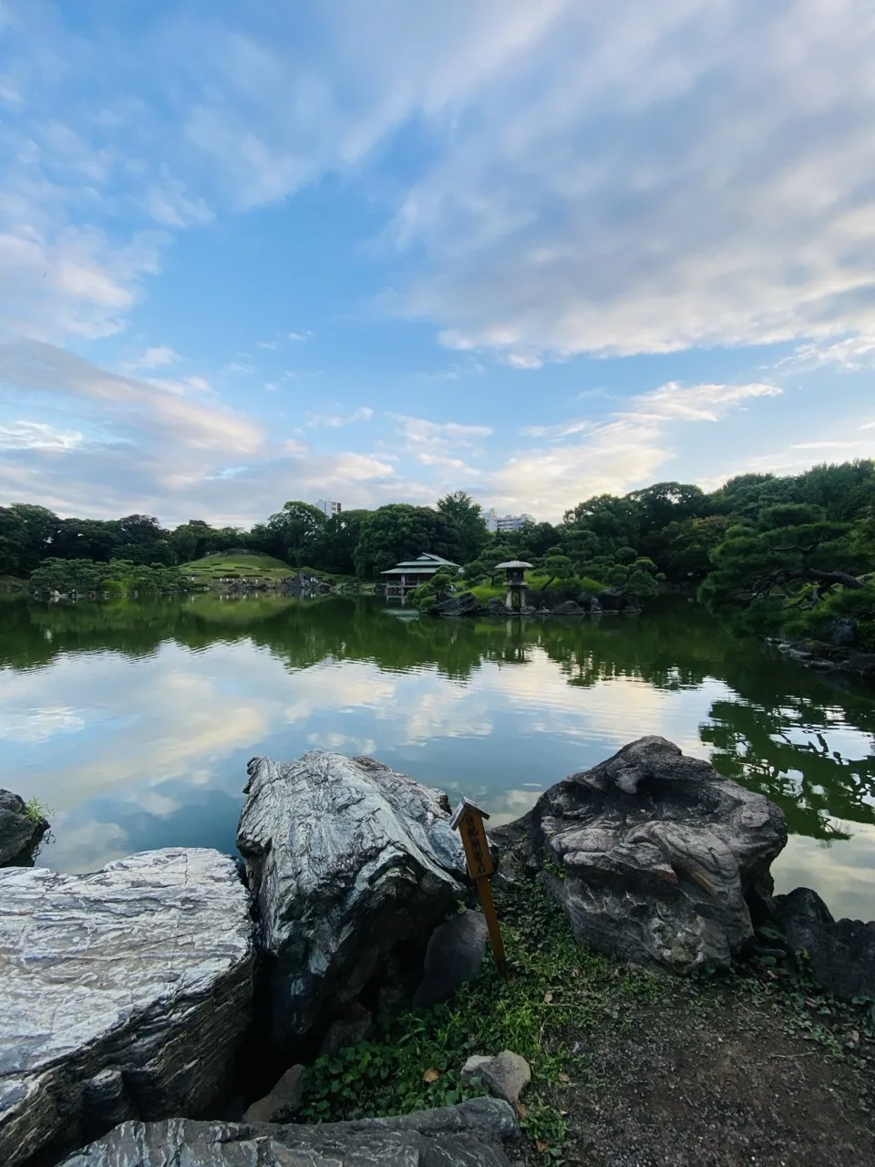画面上半分は青空と薄い雲、下半分は雲を映す穏やかな池。対岸には緑の築山と刈込み、緑青色の屋根の水亭、石灯籠が並ぶ。手前には荒々しい肌の庭石と木製の立札、足元の下草が写る