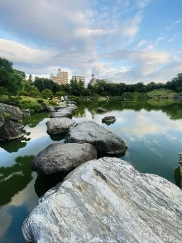 池に点々と続く大きな飛石。水面に夕方の雲が映り、奥の岸に人影と木立、ビル。