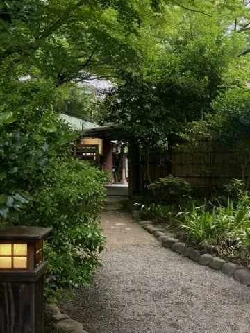 生垣と竹垣に挟まれた砂利道が奥の建物の軒先へ続く。手前左に石灯籠。軒に吊り飾り。