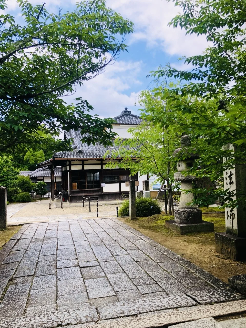 四角い石を敷き詰めた広い道が奥へ延び、突き当りに瓦屋根の堂がある。右手に石灯籠と縦書きの札、周囲は新緑の枝葉。