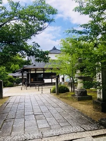 四角い石を敷き詰めた広い道が奥へ延び、突き当りに瓦屋根の堂がある。右手に石灯籠と縦書きの札、周囲は新緑の枝葉。