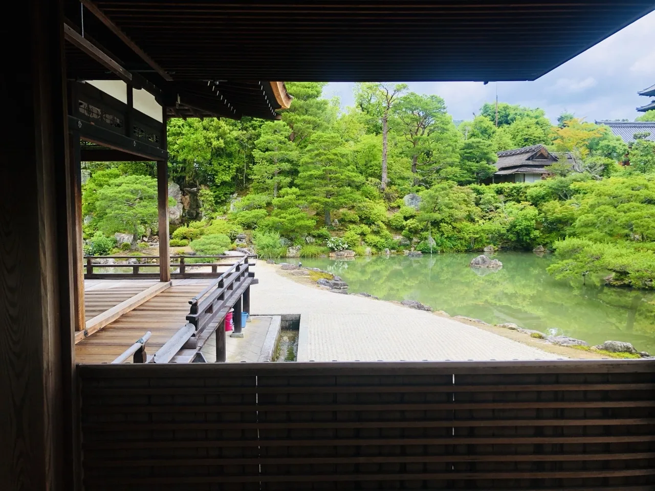 建物の暗い軒下から外を見る。手前に縁側と木の手すりが影になって横切り、その先に白砂の州と緑色の池、対岸の松と築山が明るく見える。