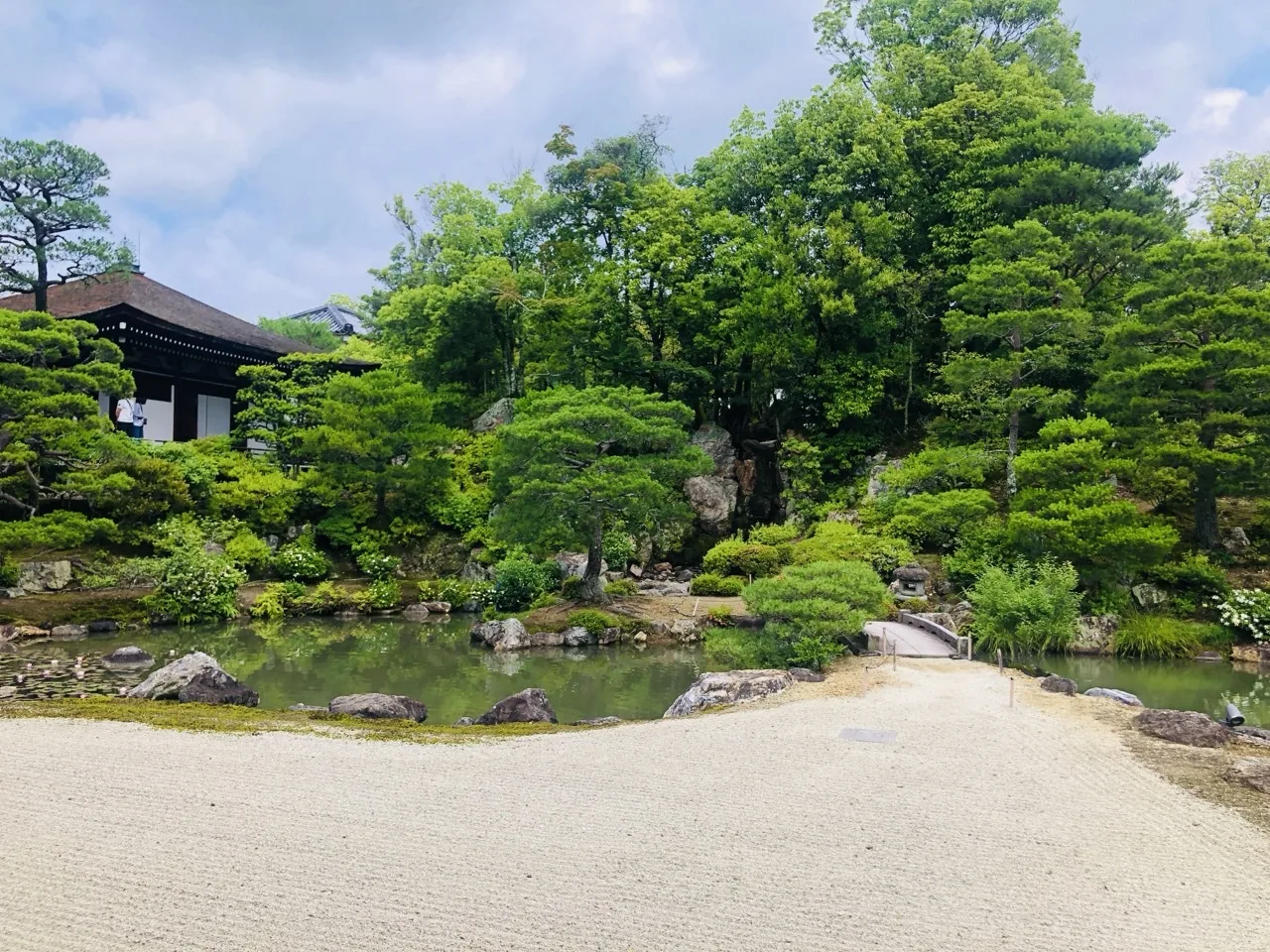 手前一面に筋を引いた白砂が広がり、その奥に浅い池と石組、刈込まれた松が並ぶ。左手に瓦屋根の建物、上は明るい曇り空。