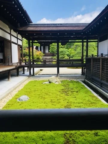 渡り廊下と建物に三方を囲まれた明るい苔の中庭。中央やや奥に白い石が一つ置かれ、左手前にもう一つ小さな石がある。奥に青空と木立。
