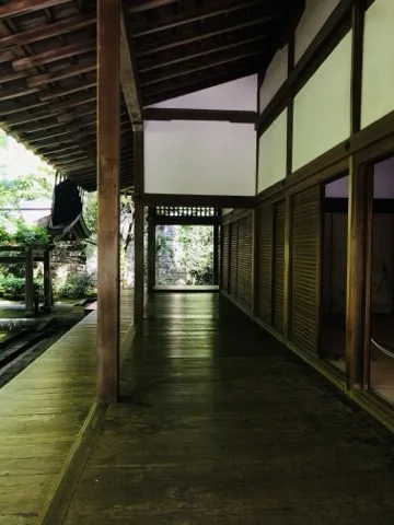 寺の広い板張りの廊下。左手は柱越しに庭、右手に障子と格子戸が続く。奥に明るい開口。